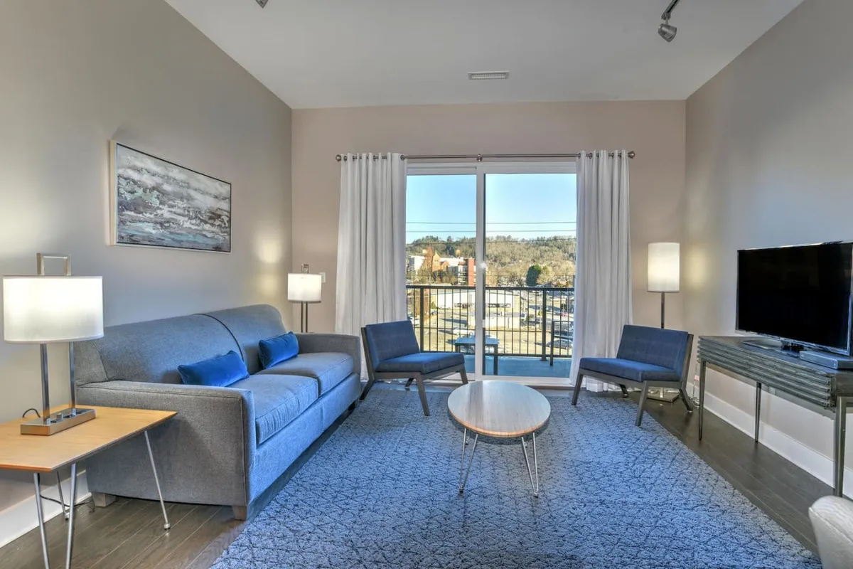 A contemporary living area with a plush sofa, coffee table, armchairs, and a TV. Large windows offer a glimpse of the view outside, and the space is arranged for comfort and social activities like movie nights or game gatherings.
