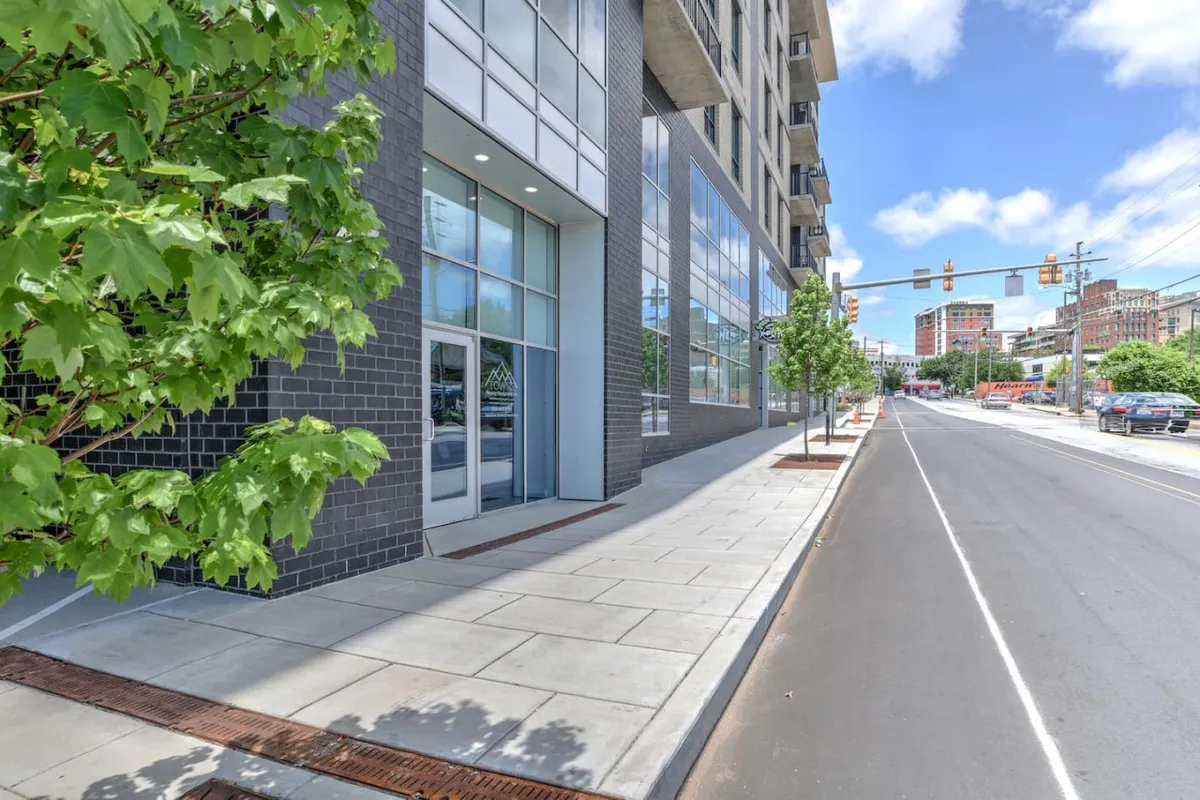 This image displays a lively streetscape featuring a modern condo building facade, sidewalk greenery, and an active street scene with traffic. People can be seen walking, biking, and socializing at nearby cafes, embodying the energy of downtown Asheville. The sidewalk is inviting and the building’s urban design blends seamlessly with its vibrant surroundings.