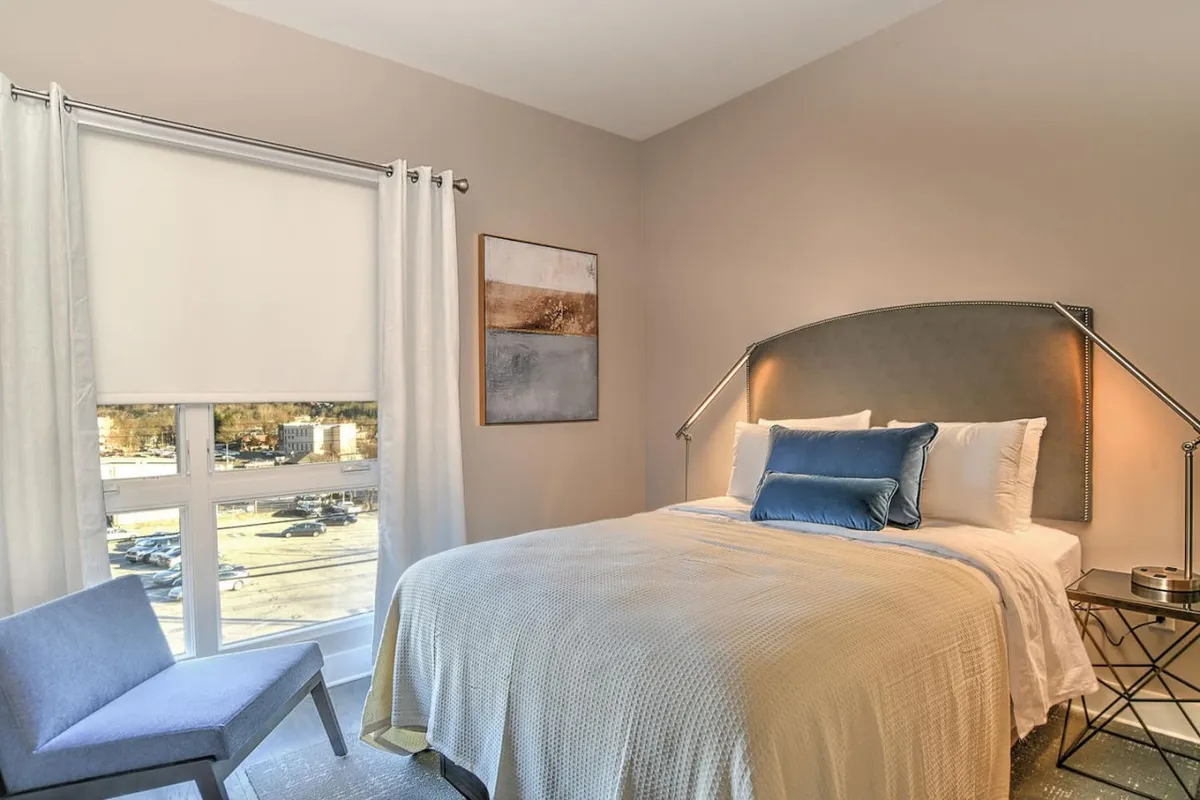 A cozy bedroom featuring a comfortable bed adorned with decorative pillows, soft lighting, and artwork on the wall. Sunlight filters through the window, creating a serene atmosphere for relaxation or reading.