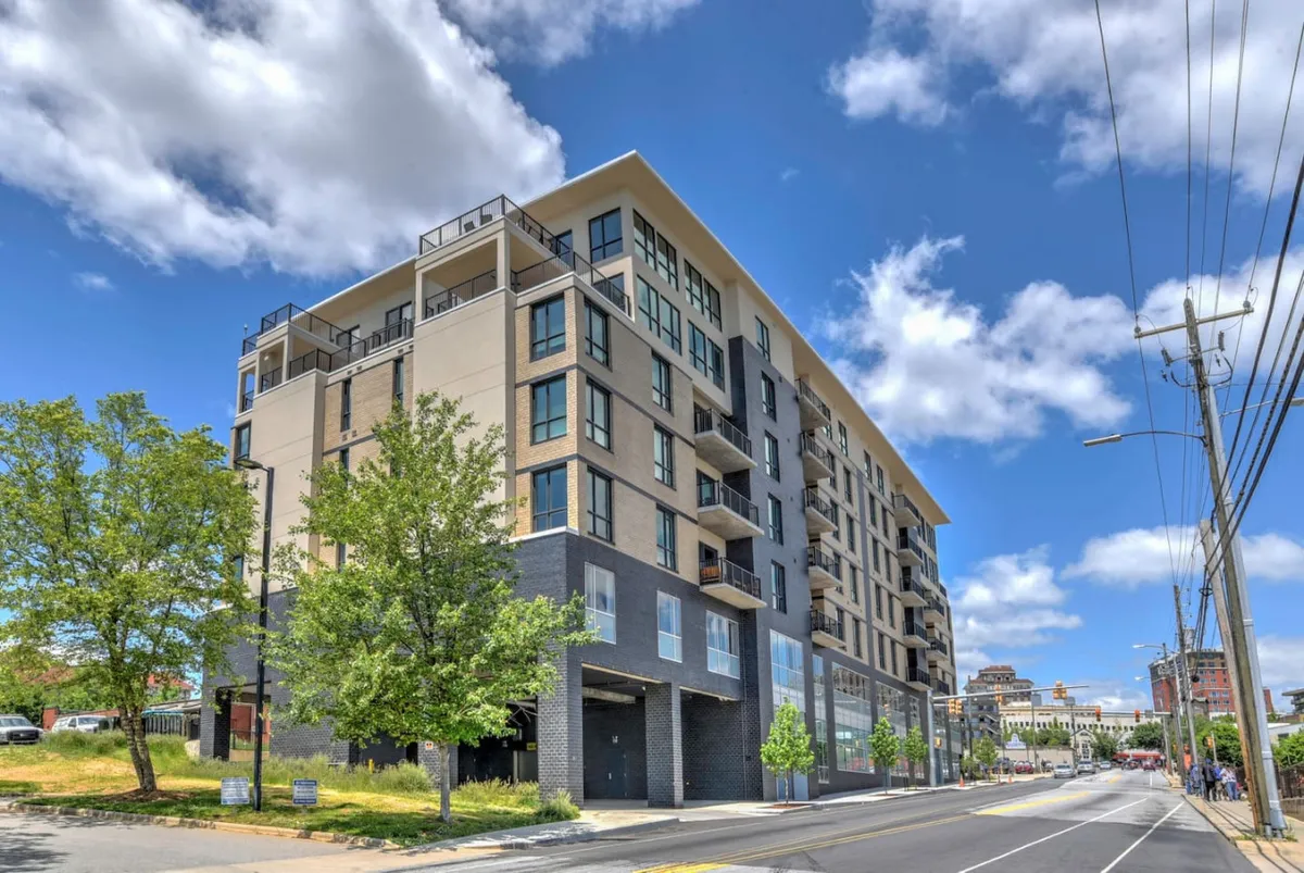 A contemporary exterior shot of the building with modern architectural design, balconies, landscaped greenery, and a street view. The lively environment suggests an urban lifestyle with secure, managed access points and proximity to city amenities.