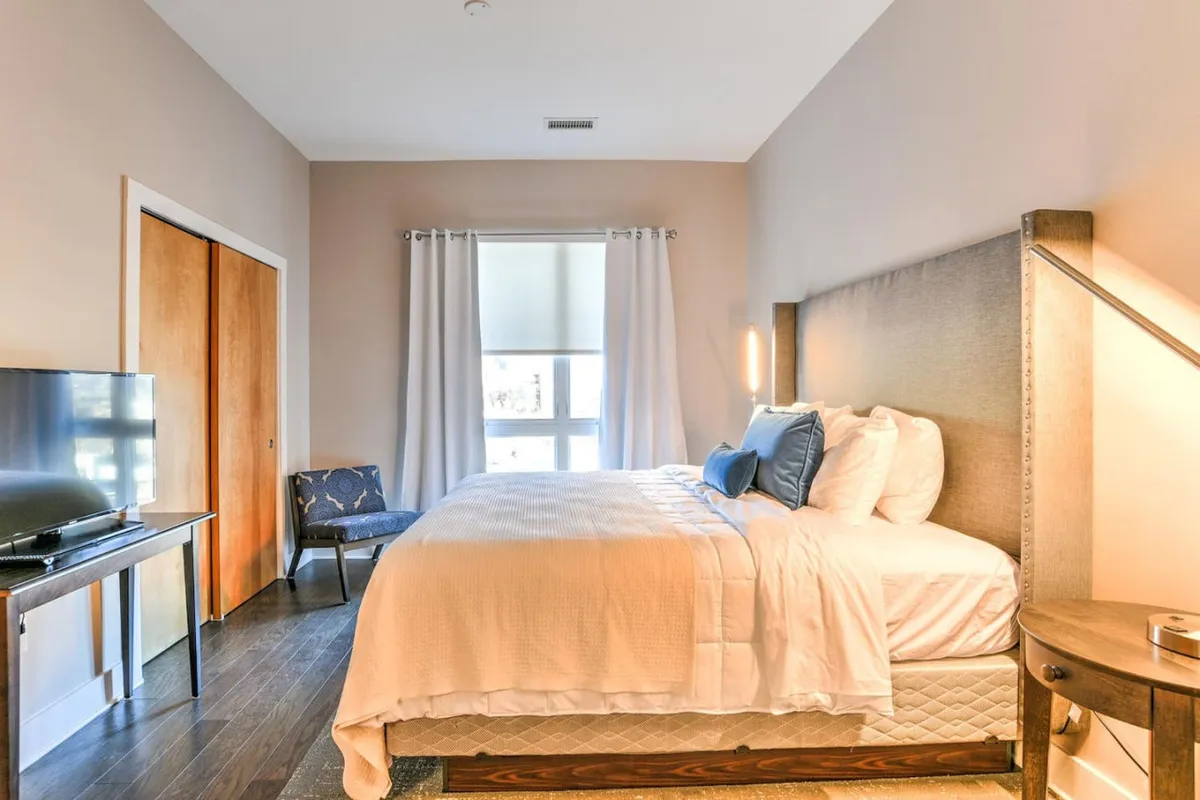A serene bedroom scene featuring a plush bed, a cozy reading chair by the window, and a television. Natural light fills the room, creating a restful and welcoming environment for both guests and their pets to settle in.